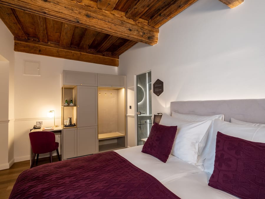 A bedroom with a wooden ceiling, a bed with white and maroon bedding, a desk with a chair, and a mirror.