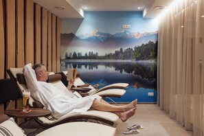 A man relaxing in a sun lounger with a book and a glass of beer, a lake mural, and wooden paneling on the walls. - Das Glemm Hotel (Saalbach-Hinterglemm)