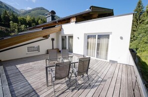 Una terraza de madera con mobiliario exterior, un edificio blanco con amplios ventanales y montañas al fondo.