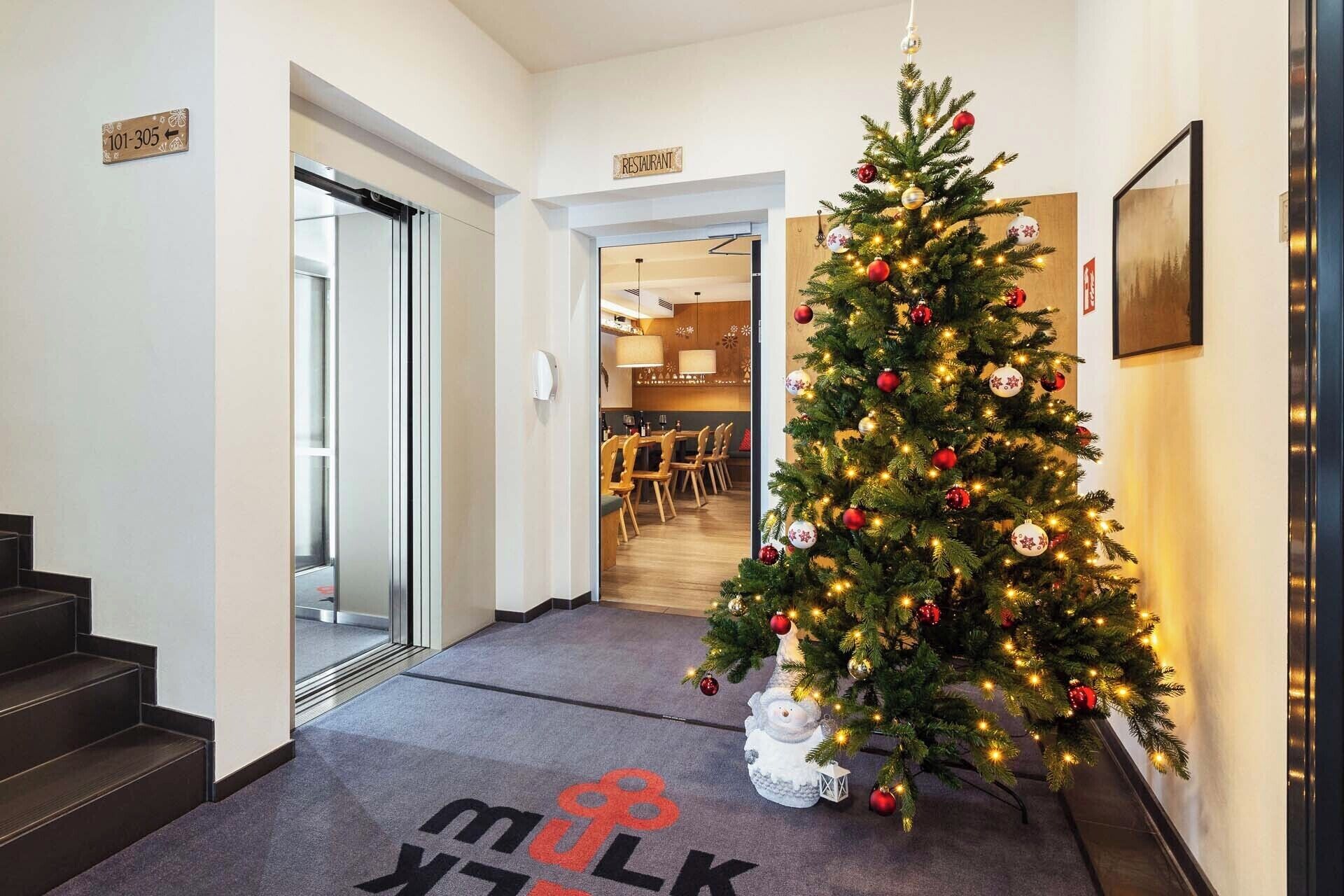 A hotel hallway with a Christmas tree, a sign indicating a restaurant, and a carpet with a logo.
