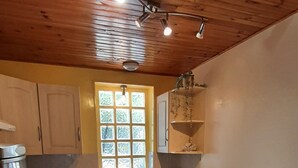 A kitchen with wooden ceiling, white cabinets, a window, a sink, a stove, and a table with a chair.