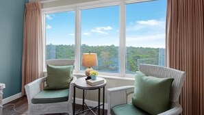 Ein Zimmer mit zwei Korbstühlen, einem kleinen Tisch mit Lampe und Blick auf einen Wald durch große Fenster.