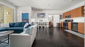 Un salon moderne avec un canapé d’angle, un coin repas avec une table et des chaises blanches, et une cuisine dotée de meubles en bois et d’appareils électroménagers en acier inoxydable.