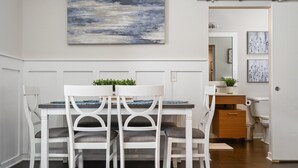 Un coin repas avec une table et des chaises blanches, un tableau au mur et une vue sur la cuisine.