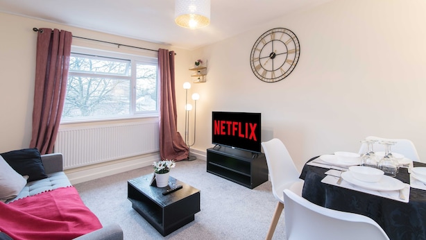 A living room with a sofa, a coffee table, a TV displaying 'NETFLIX', a dining table set with plates and glasses, and a large wall clock. - Coventry- Jenner Pet Friendly 2 Bedroom Apartment (Coventry)