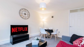 A living room with a TV displaying 'NETFLIX', a dining area with a table and chairs, a lamp, and a door. - Coventry- Jenner Pet Friendly 2 Bedroom Apartment (Coventry)