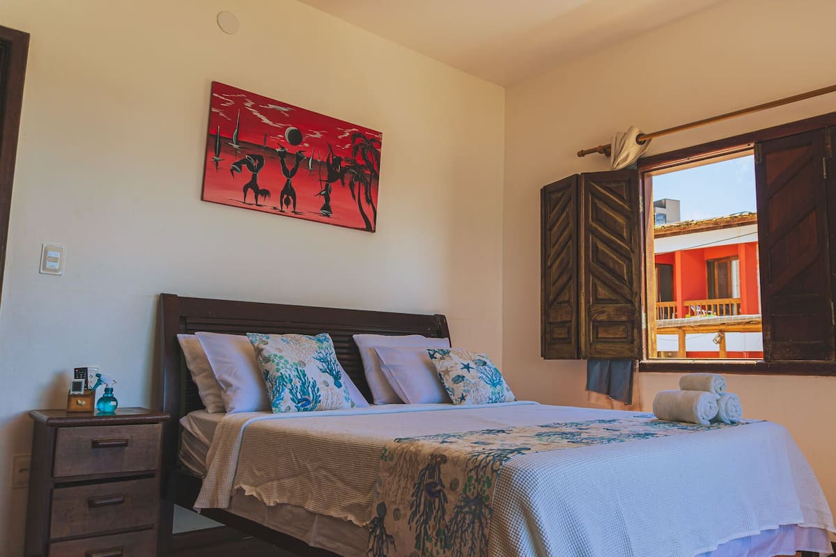 Quarto com cama, mesa de cabeceira, pintura e janela com cortinas.