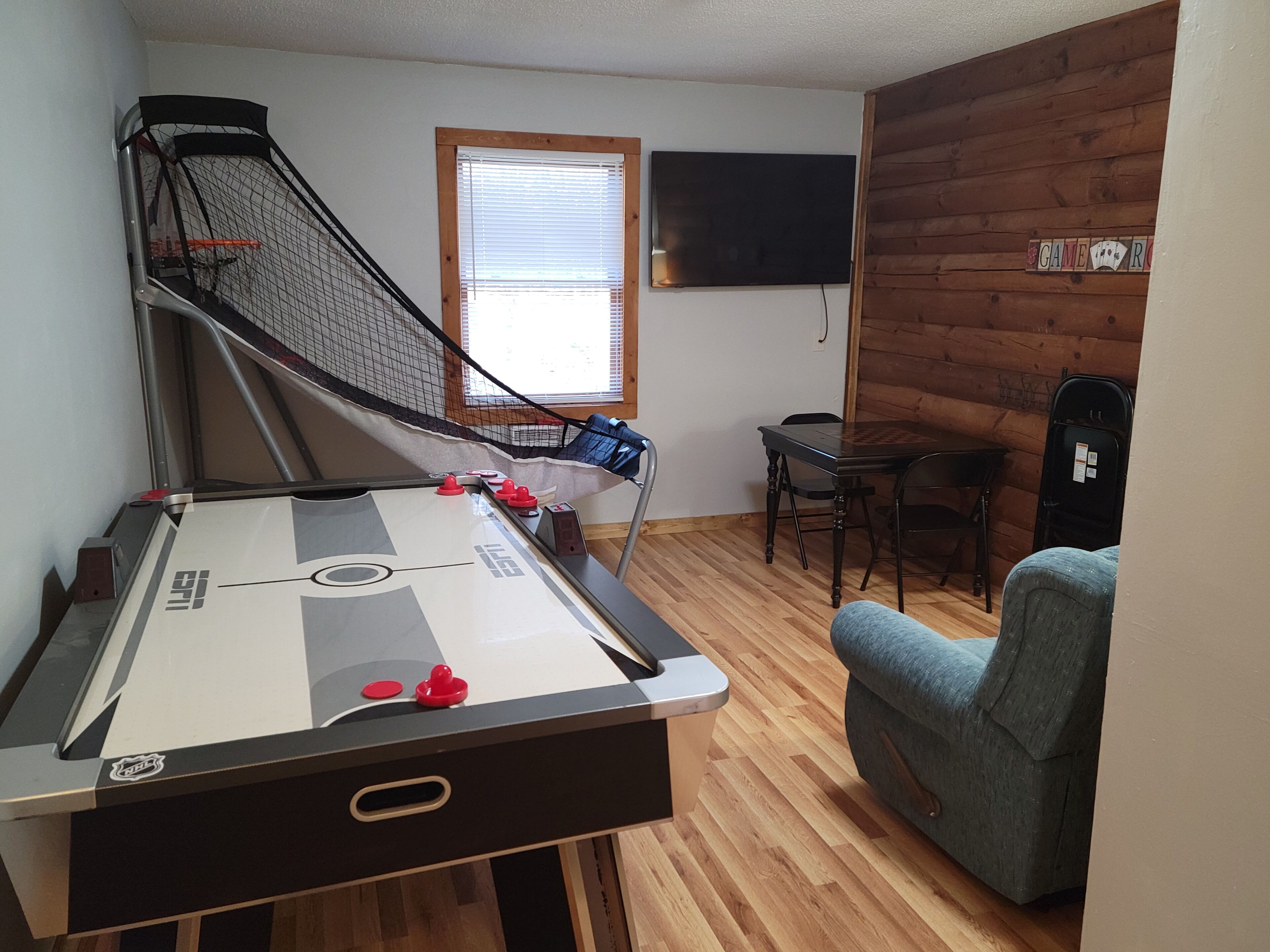 A pool table with a shuffleboard, a blue armchair, a wooden table, and a television mounted on the wall.