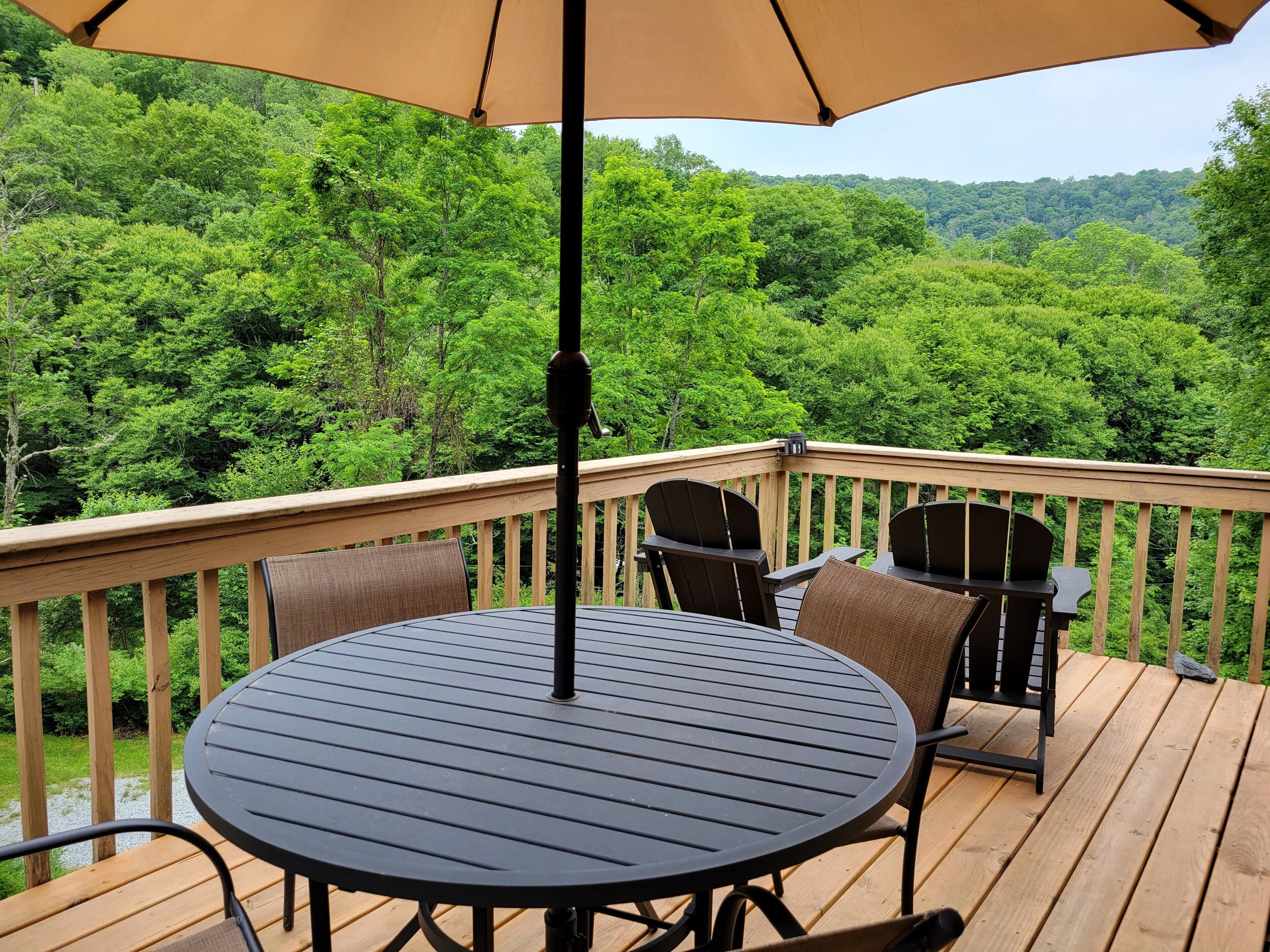 Une terrasse dotée d’une table ronde et de chaises, entourée d’une rambarde en bois, et offrant une vue sur une forêt dense.