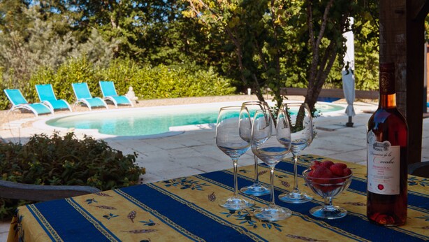 Un tavolo a bordo piscina con bicchieri da vino e una bottiglia, circondato da poltroncine blu e una piscina.