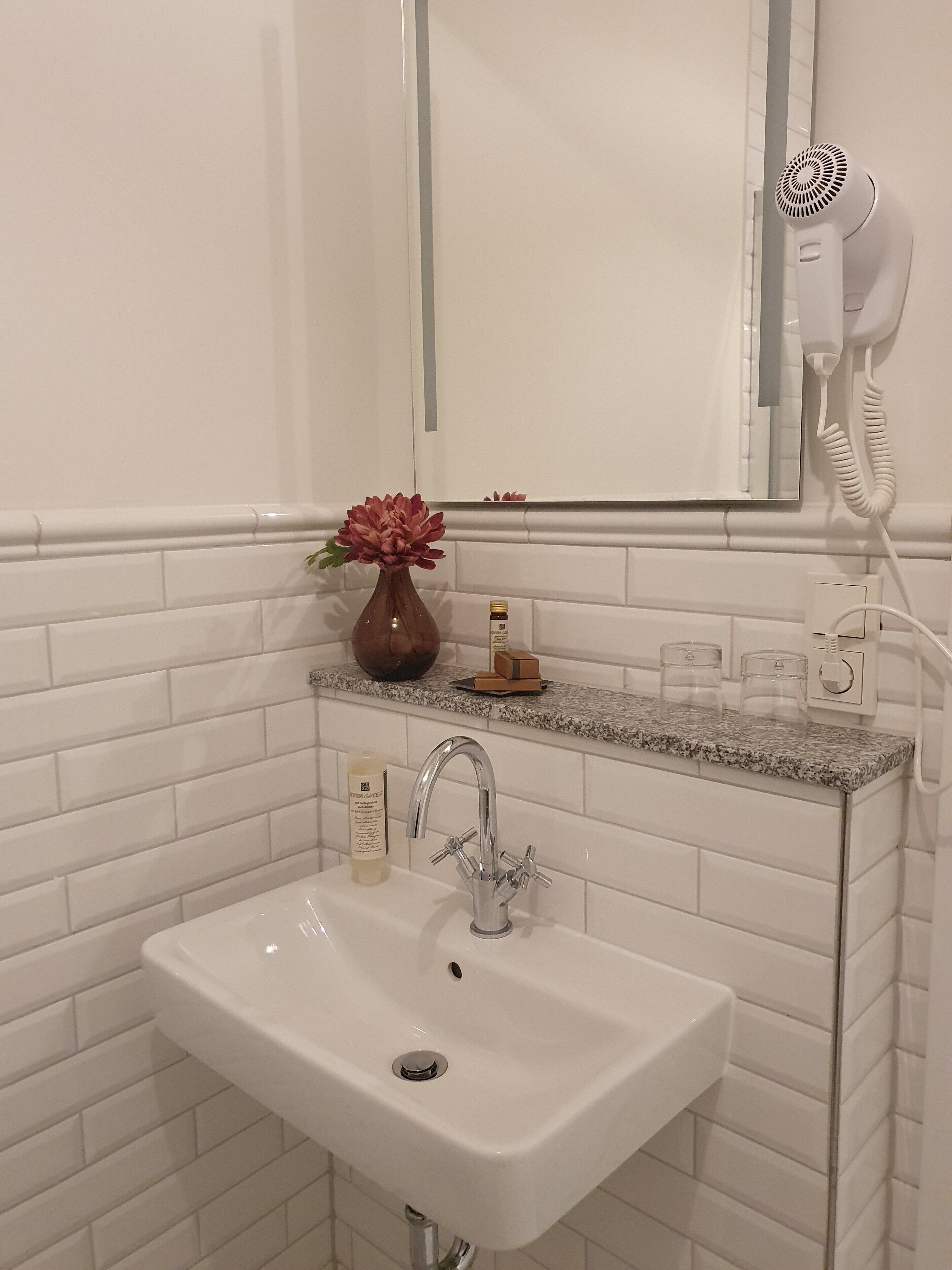 Un baño con lavamanos blanco, espejo, secador de pelo y un florero con flores.