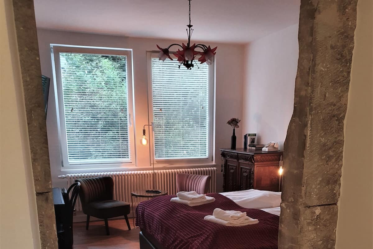 Un dormitorio con una cama grande, una silla, un escritorio y dos ventanas con persianas.