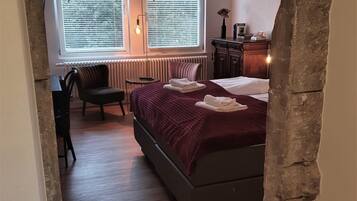 Une chambre avec un grand lit, une chaise, un bureau et deux fenêtres équipées de stores.