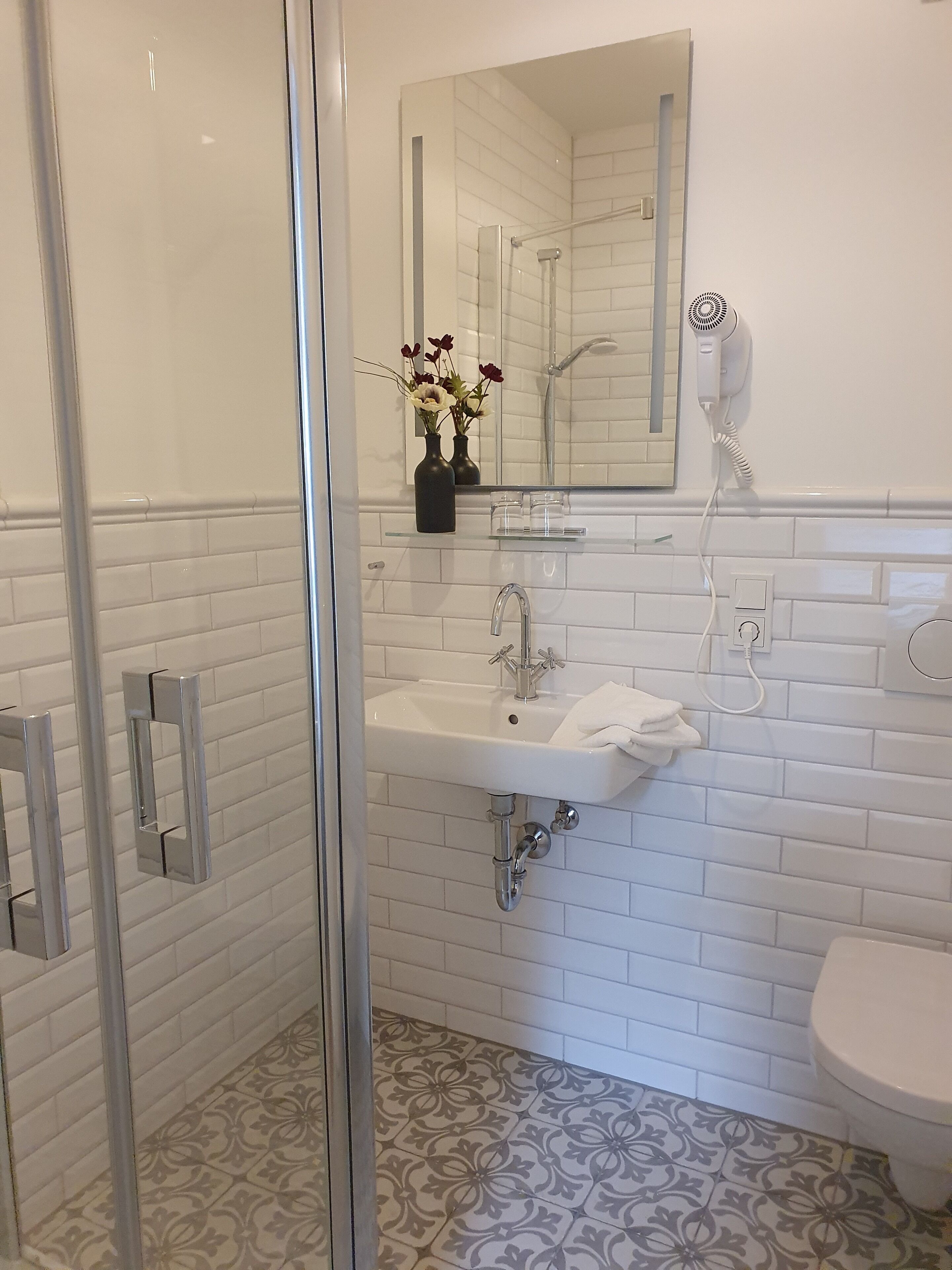 A modern bathroom with a glass shower enclosure, a white sink with a mirror above, and patterned floor tiles.