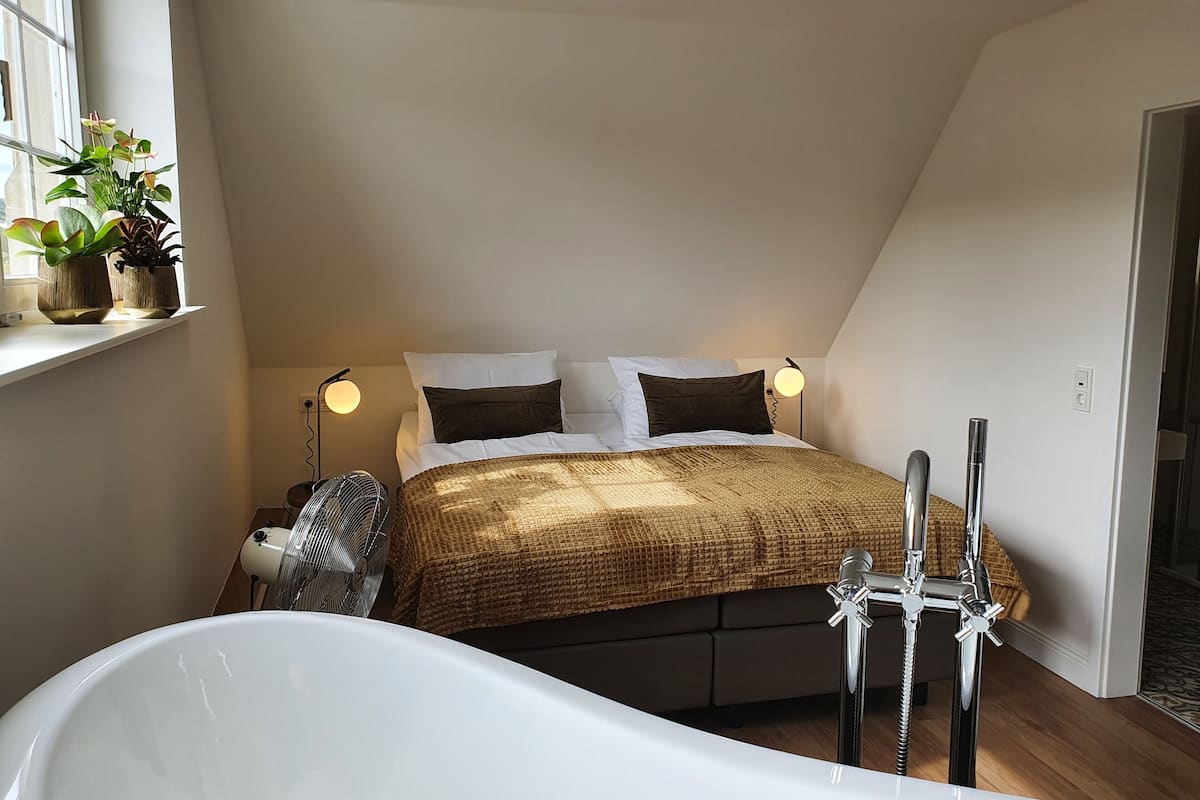 Un dormitorio con cama, dos lámparas de noche, una ventana con plantas y una bañera.