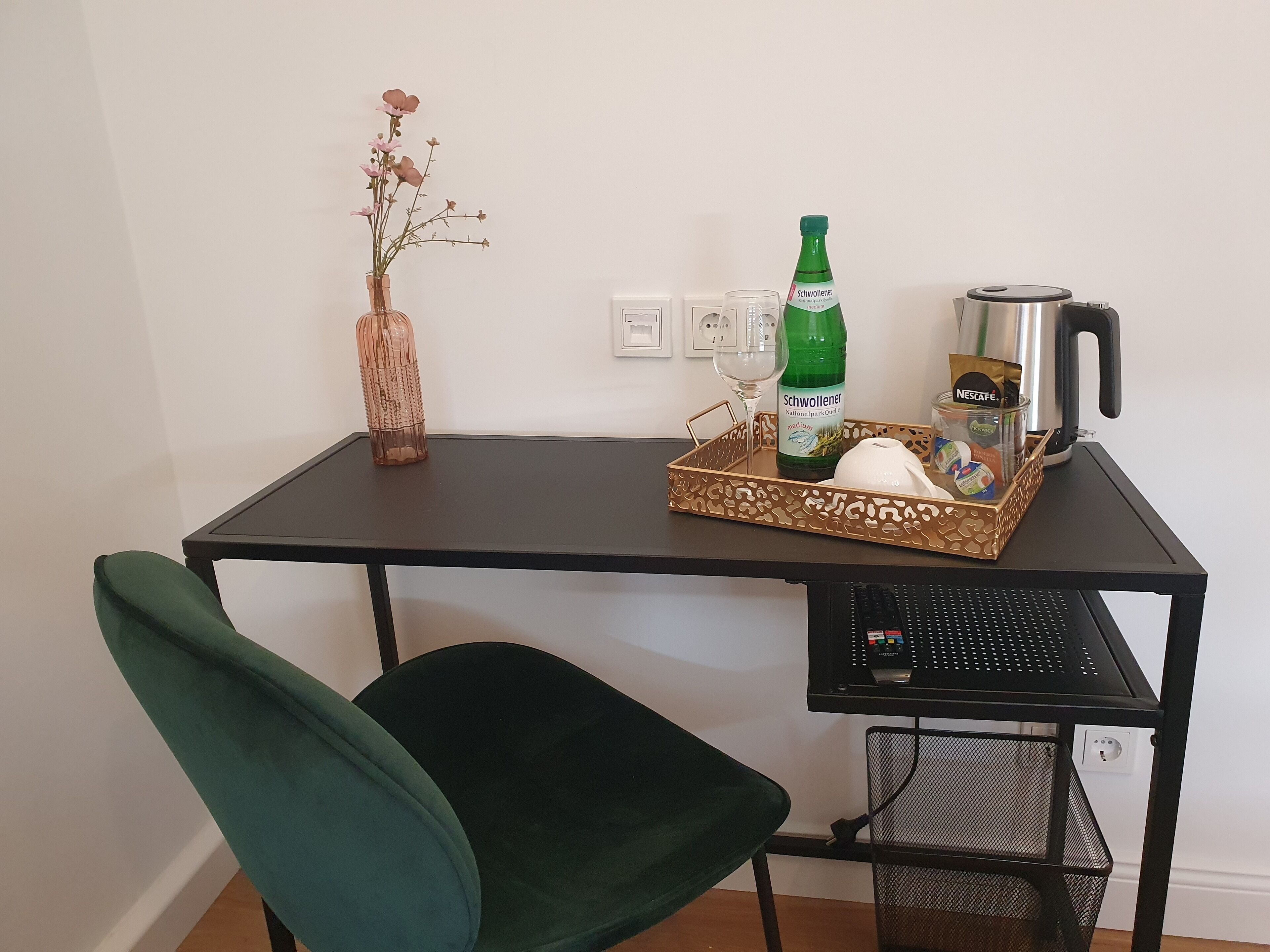 Un escritorio pequeño y oscuro con una silla verde, un jarrón con flores rosas, una botella de Schweiller, un vaso, una cafetera Nescafé y una cesta con un objeto blanco.