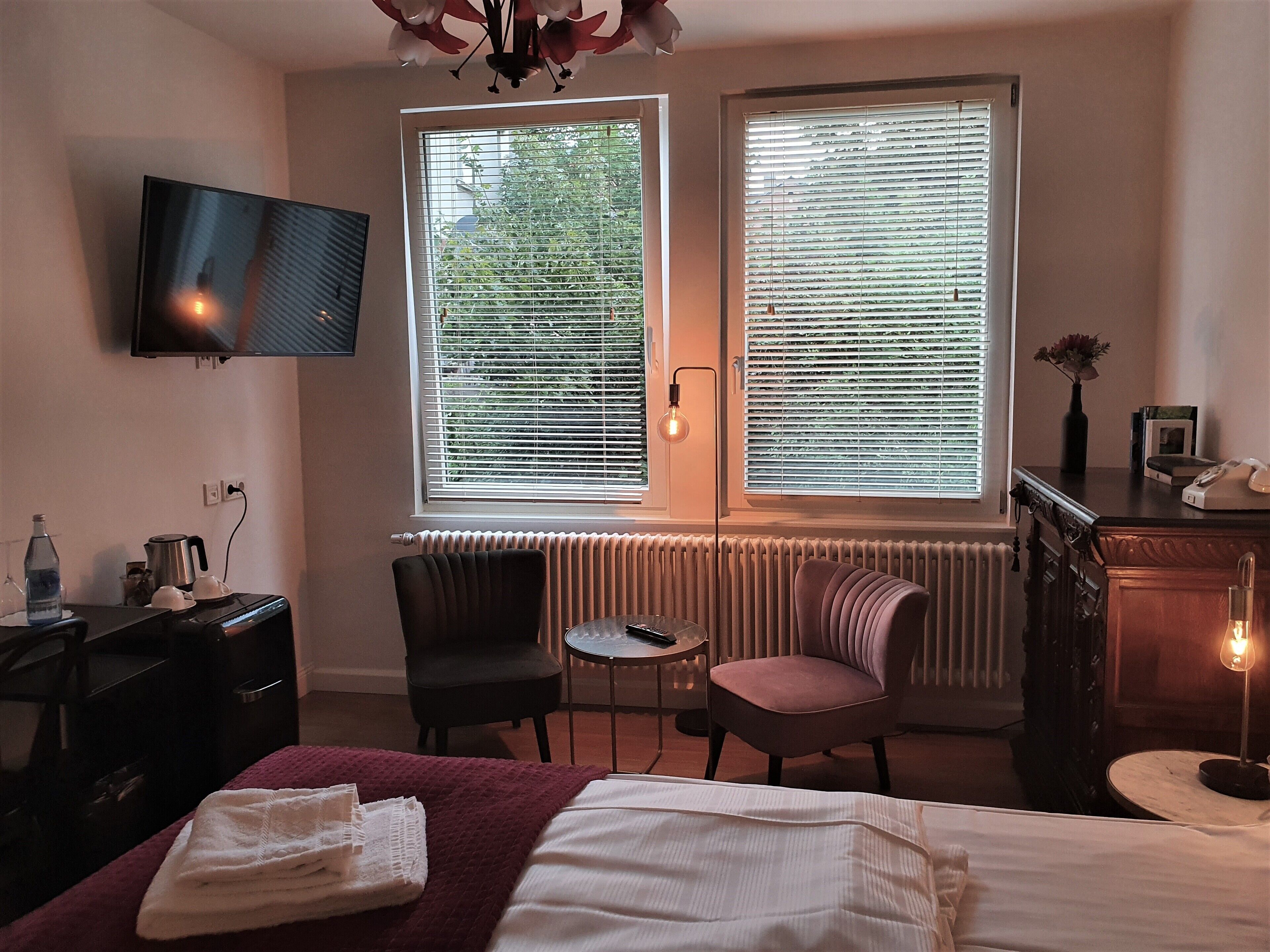 A hotel room with a bed, a desk, a chair, a small table, and two windows with blinds.