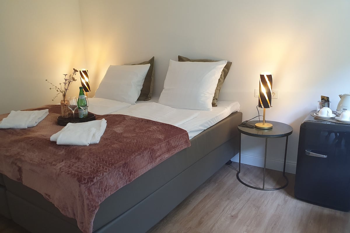 Una cama bien hecha con ropa de cama blanca y marrón, una mesita de noche con una lámpara, y una pequeña mesa con una tetera y tazas.