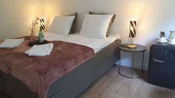 Cama bem arrumada com roupa de cama branca e castanha, mesa de cabeceira com abajur e uma pequena mesa com chaleira e chávenas.