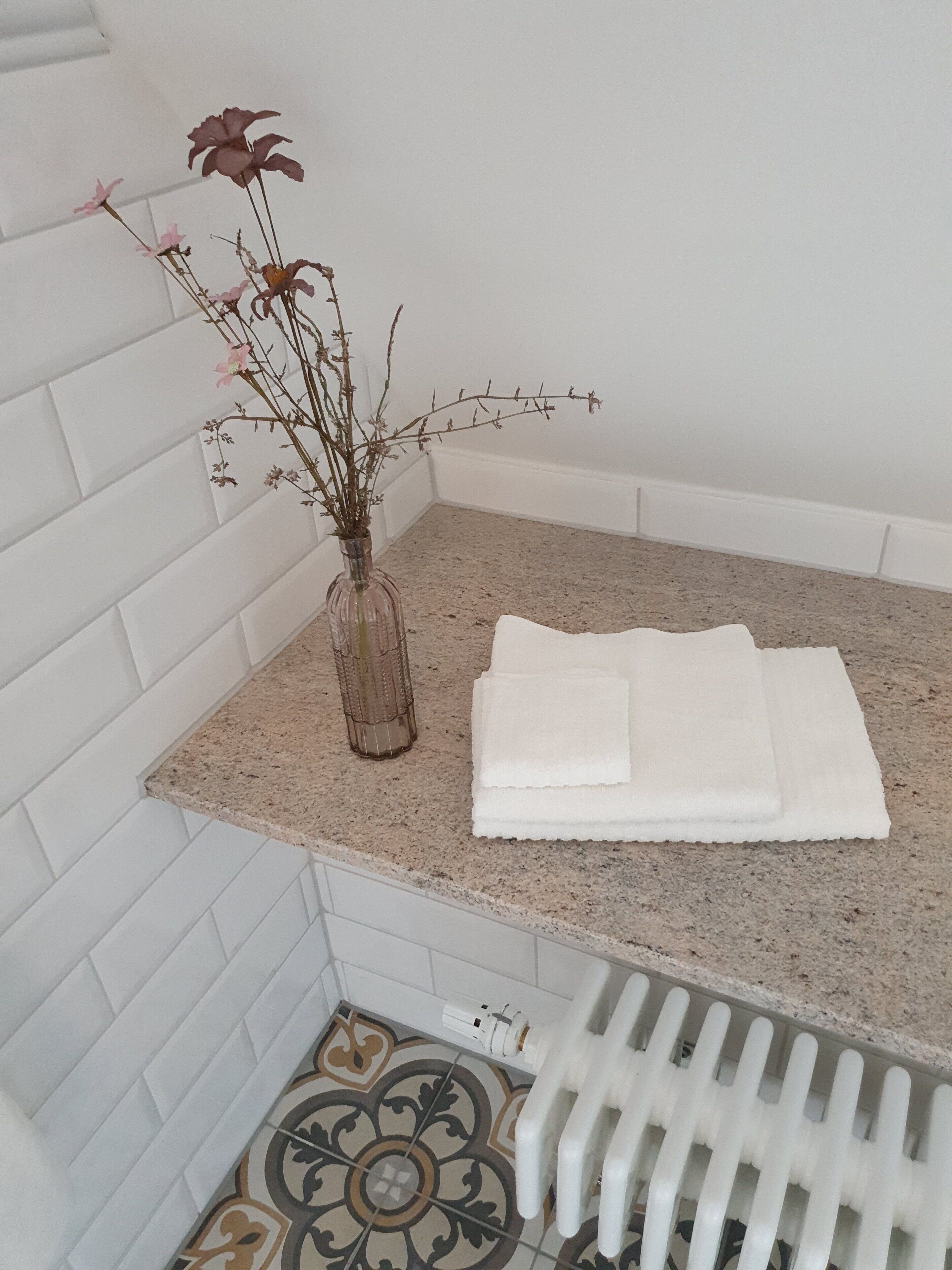 A bathroom with white tiled walls, a granite countertop, a decorative vase with dried flowers, and a folded white towel.