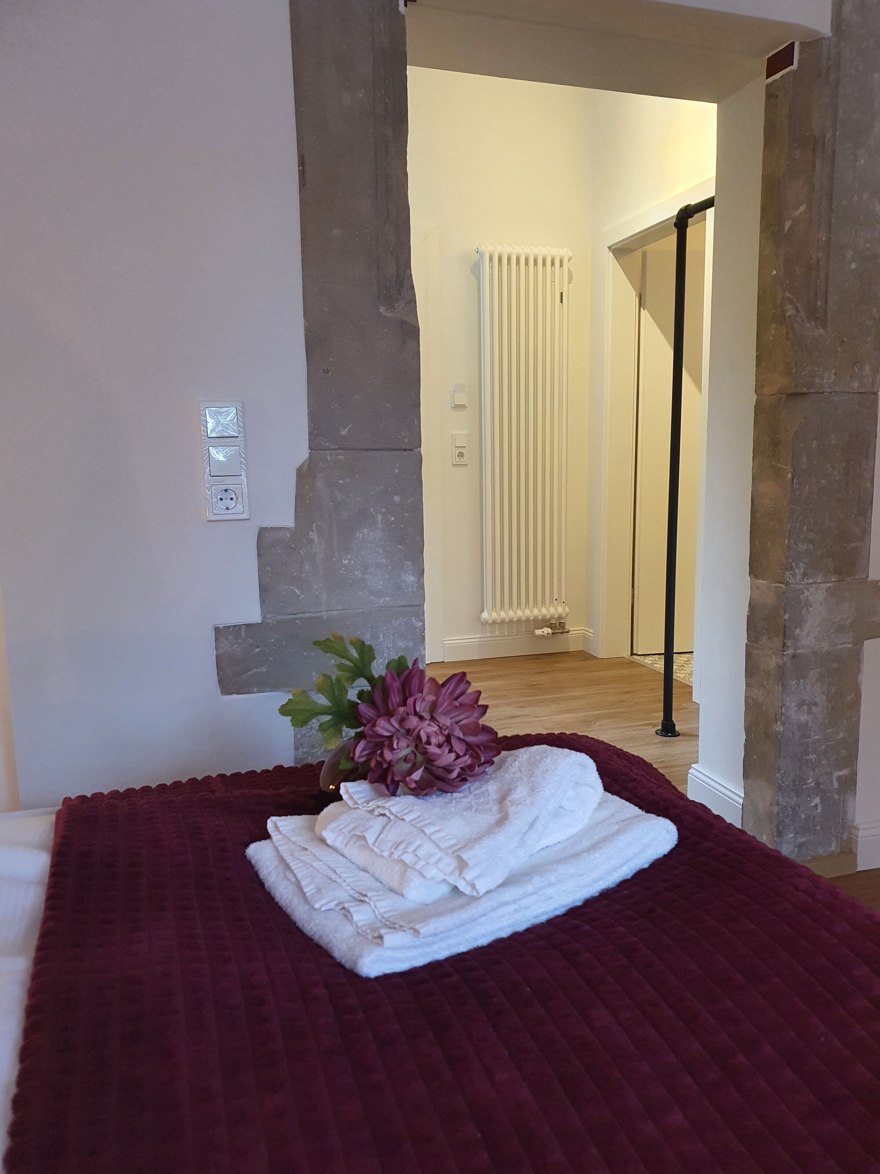 A neatly made bed with a folded white towel and a decorative plant in a room with stone walls and a radiator in the background.