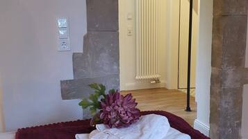 A neatly made bed with a folded white towel and a decorative plant in a room with stone walls and a radiator in the background.