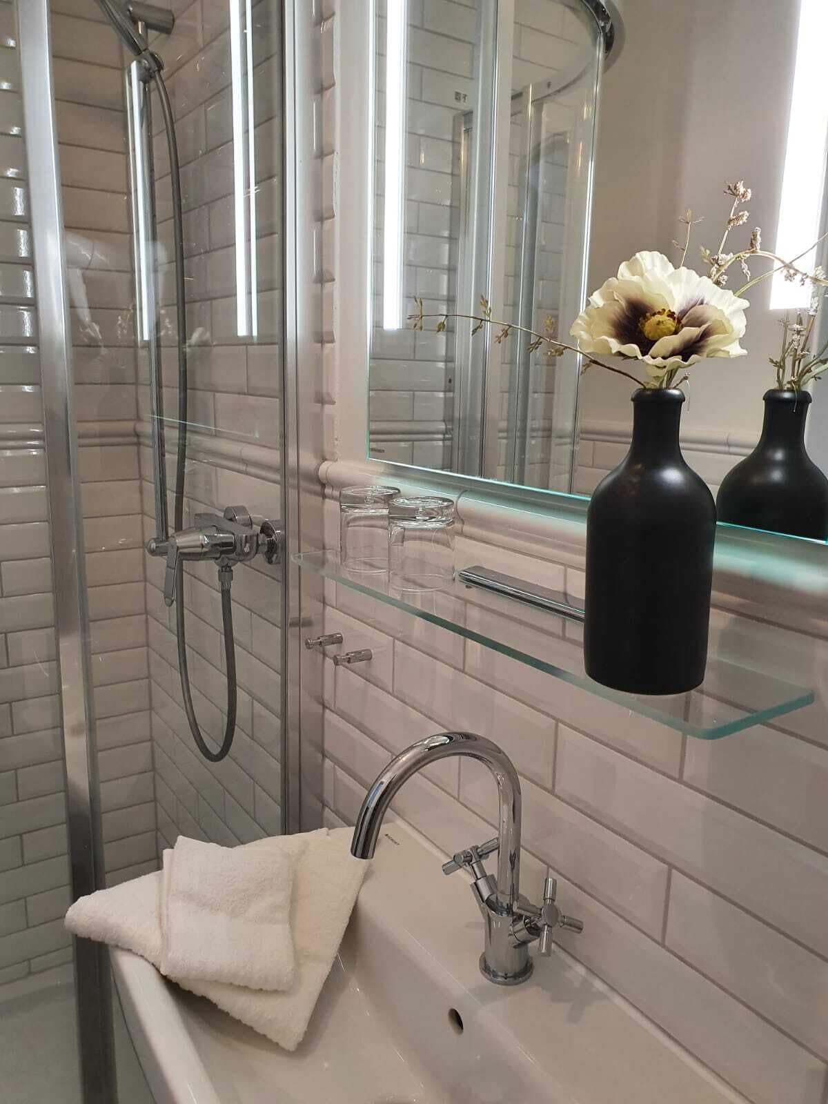 Un baño moderno con cabina de ducha de vidrio, un lavamanos blanco con un solo grifo y un espejo sobre el lavamanos. Hay dos jarrones negros con flores secas en un estante de vidrio.