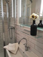 Un baño moderno con cabina de ducha de vidrio, un lavamanos blanco con un solo grifo y un espejo sobre el lavamanos. Hay dos jarrones negros con flores secas en un estante de vidrio.