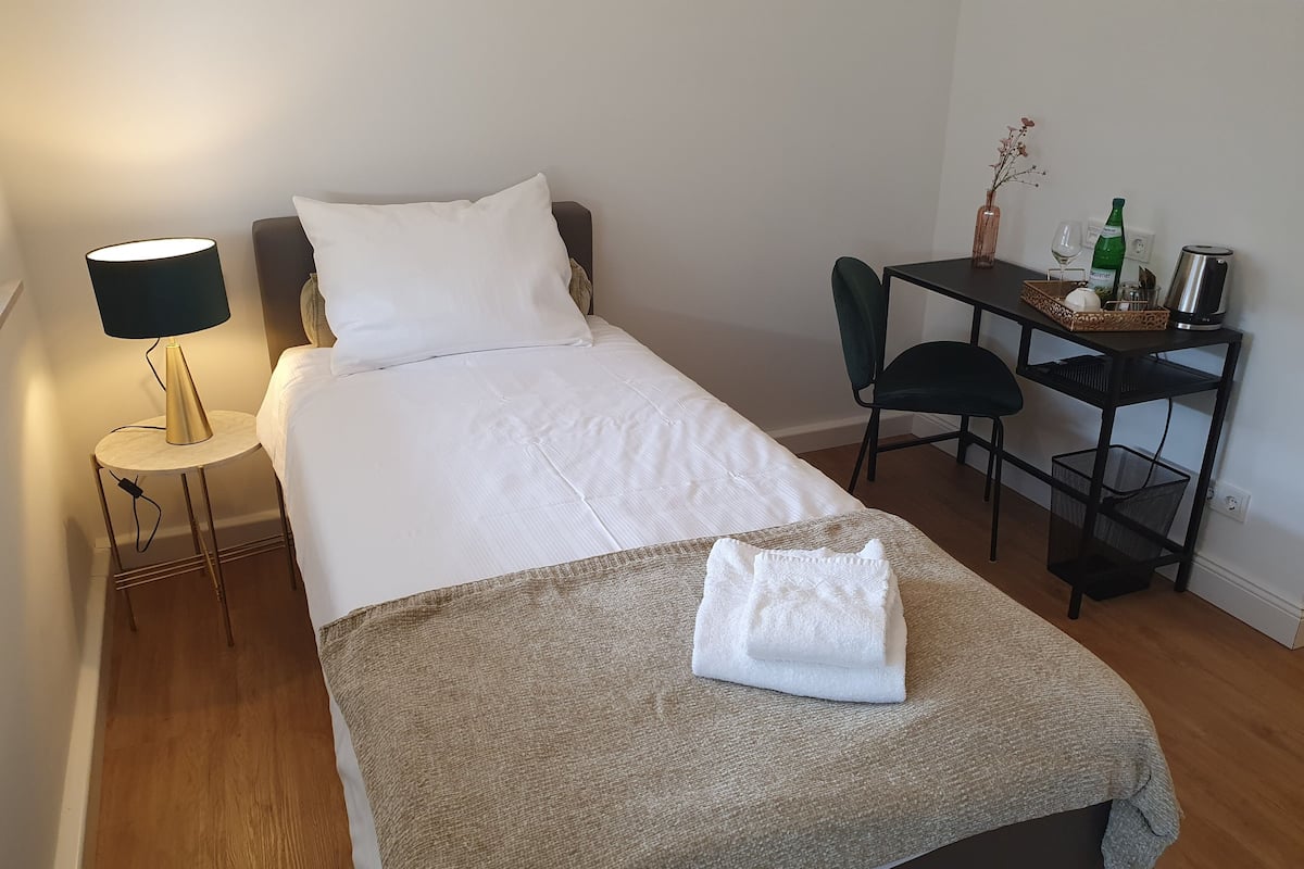 Una cama bien hecha con un edredón blanco y una manta color beige, una mesita redonda con una lámpara, un escritorio con una silla, y un jarrón con flores.