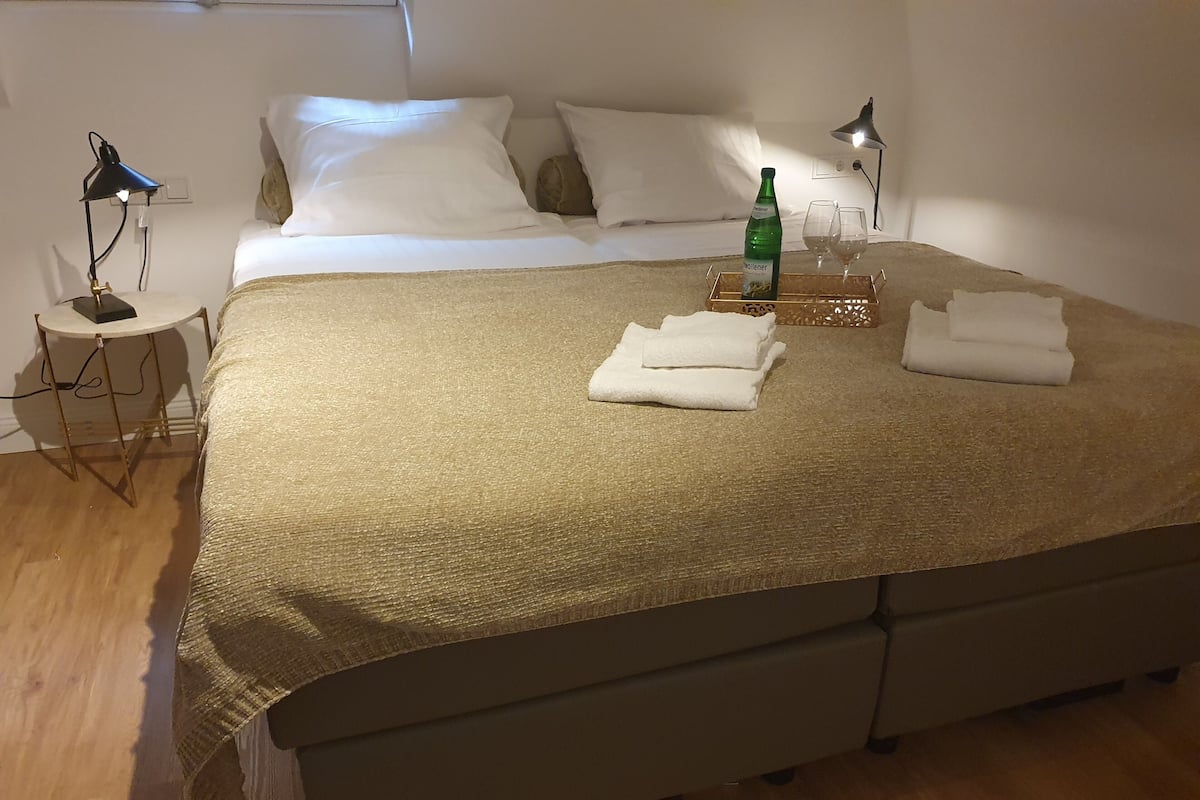 Una cama bien hecha con una manta color beige, dos lámparas de noche, una botella de agua y toallas dobladas sobre una mesita de noche.