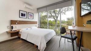 A modern hotel room with a large bed, a desk, and a chair. There is a window with a view of trees and a balcony. - Hotel Park 85 (Cali)