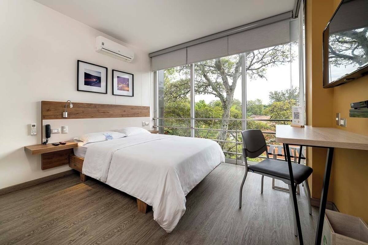A modern hotel room with a large bed, a desk, and a chair. There is a window with a view of trees and a balcony.