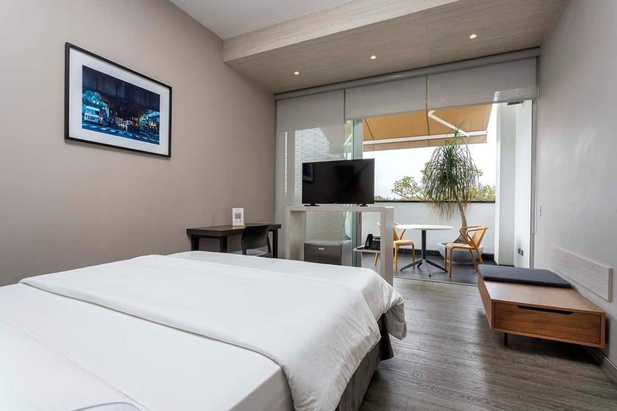 A modern hotel room with a large bed, a desk, a chair, a television, and a window with a view of the city.