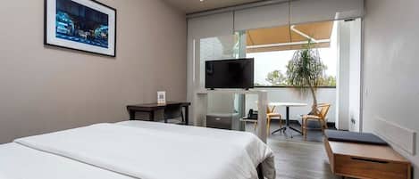 A modern hotel room with a large bed, a desk, a chair, a television, and a window with a view of the city.