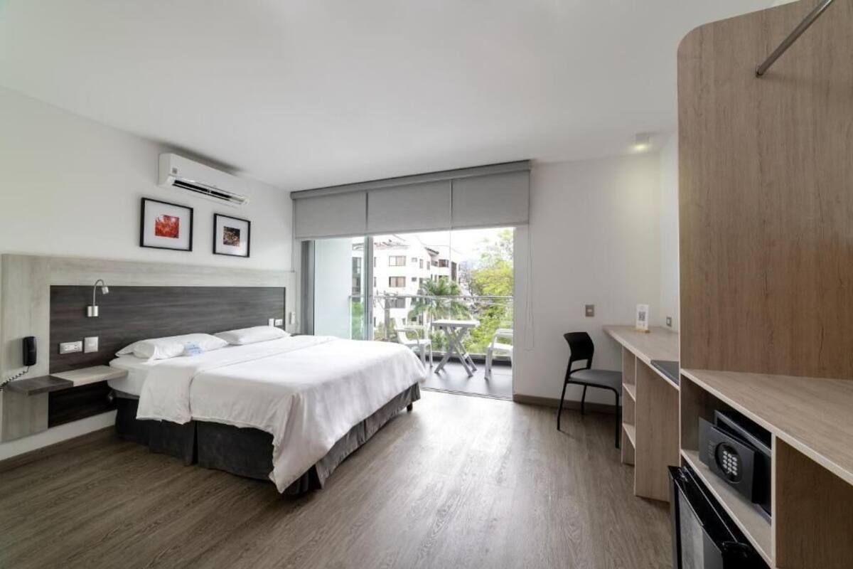 A modern bedroom with a large bed, a desk, and a balcony with a view of buildings.