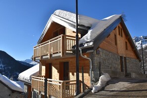 A wooden chalet with a balcony, stone walls, and a snow-covered roof.