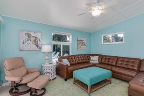 A living room with a brown leather sectional sofa, a beige armchair, a teal ottoman, a white side table with a lamp, and artwork on the walls.