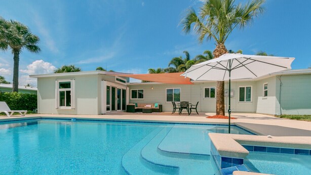 Piscina com água azul cristalina, cercada por uma casa com alpendre e mobiliário de exterior.