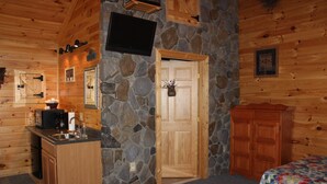 A cozy cabin interior with a stone wall, wooden walls, a kitchenette, a bed, and a television mounted on the wall.