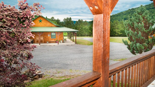 A lodge with a green roof, wooden exterior, and a star emblem. It features a covered porch with chairs and a table, surrounded by a gravel driveway and a forested hillside.