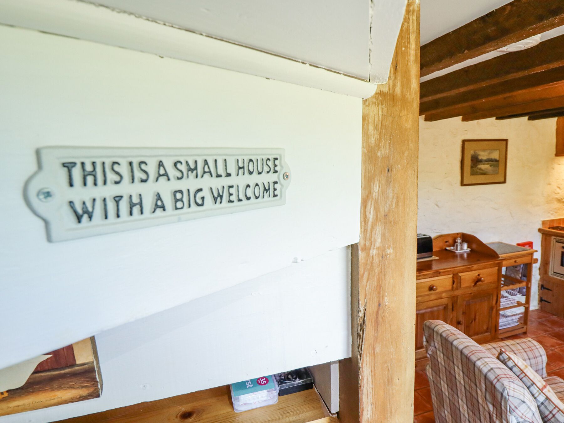 Een klein interieur met een houten tafel, een geruite bank en een bord op de muur waarop staat: “THIS IS A SMALL HOUSE WITH A BIG WELCOME”.