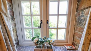 A window with a white frame and two handles, a small wooden box with flowers on the sill, and a brick wall. - Old Blacksmiths Cottage (Market Rasen)