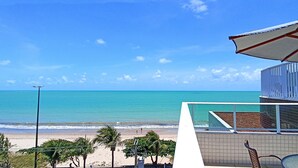 Vista de la playa con cielo despejado, arena dorada y un edificio con balcón frente al océano.