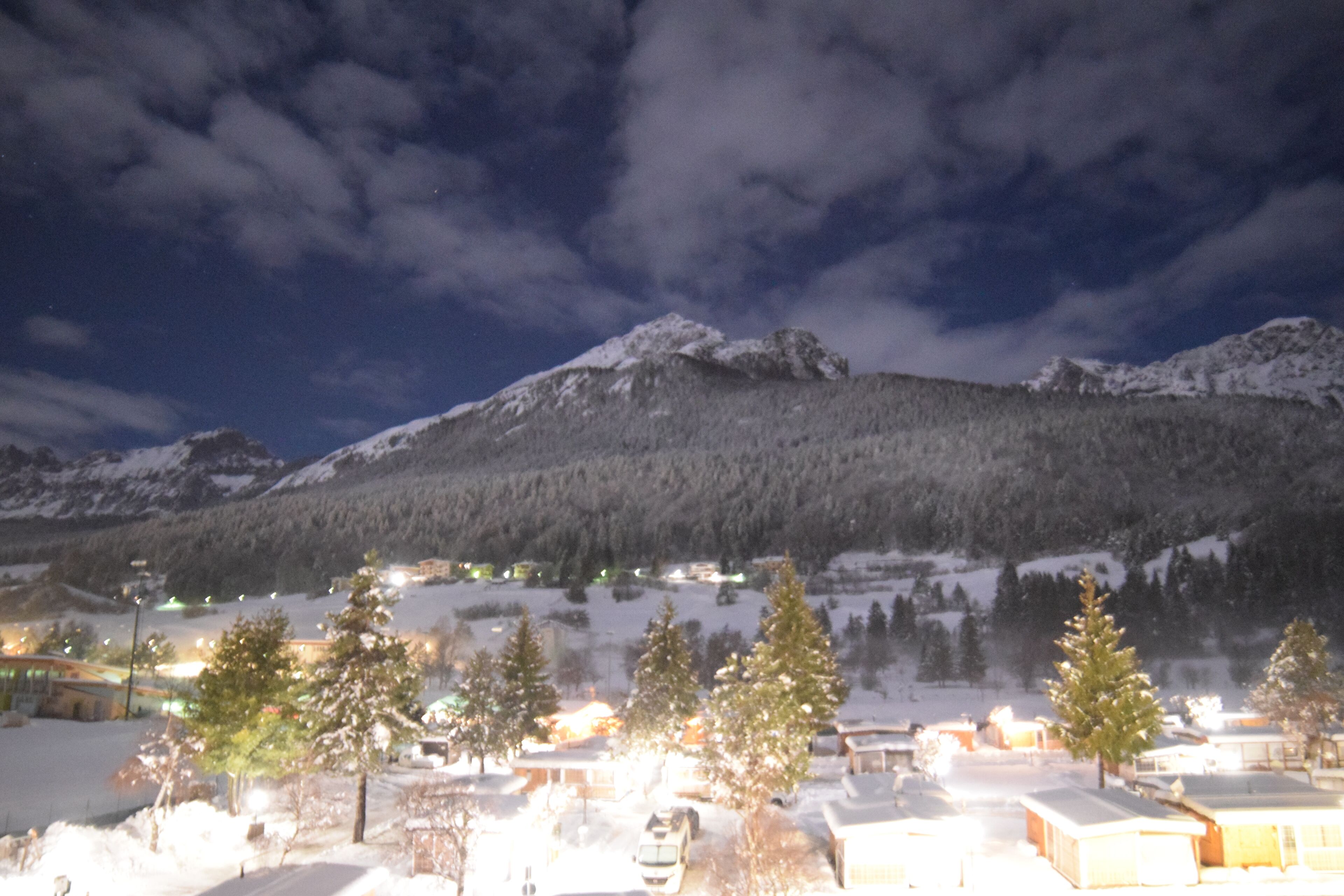 Catena montuosa innevata di notte con edifici illuminati in primo piano.