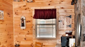 L’intérieur d’un chalet en bois comprend une table à manger, deux chaises, un lustre, une fenêtre avec des stores, un évier et une poubelle.