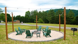 Un espace extérieur circulaire aménagé avec des chaises Adirondack vertes et un foyer central, le tout entouré d’un anneau de gravier et de poteaux en bois.