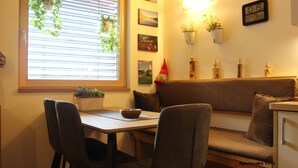 Een gezellige woonkamer met een houten tafel, twee stoelen, een bank en een raam met luxaflex. Er zijn planten aan de muur en een klok.