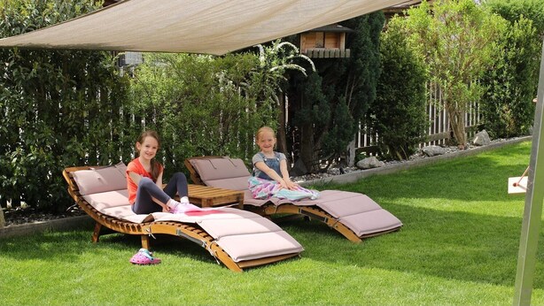 Twee kinderen zitten op ligstoelen in een tuin met een houten terras en een schaduwrijk gedeelte.