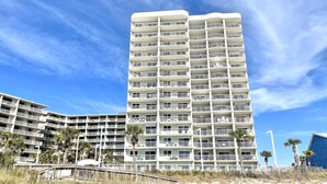 Un hôtel à plusieurs étages doté de balcons, situé près d’une plage bordée de palmiers et d’une promenade en bord de mer.