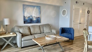 Un salon avec un canapé gris, un fauteuil bleu, une table basse en bois, une lampe et un tableau représentant des voiliers.